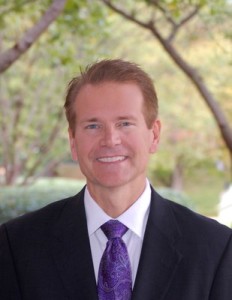 Headshot of Budenz in a suit and purple tie. The setting is outdoors.