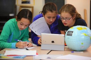 Three girls are at a classroom desk with papers and an iPad in front of them. They are working on a task together. There is also a small inflatable globe on the desk.
