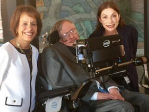 Two women posing next to Stephen Hawking who is in a wheel chair