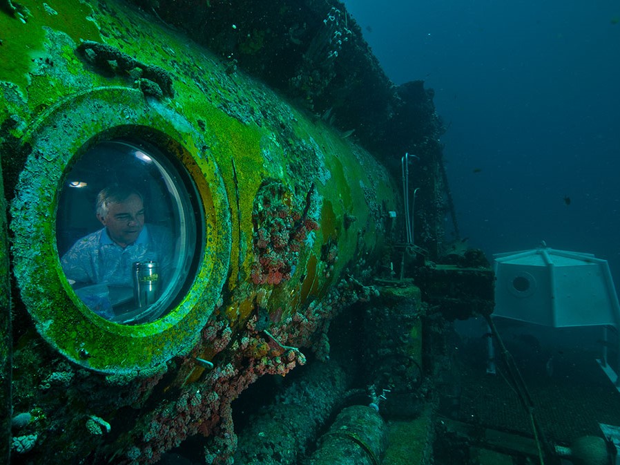Chris Martens looks out of the round viewport on underwater Aquarius Reef Base, which has green algae growing on it.