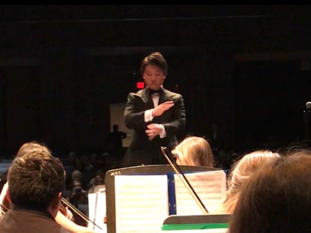 View of orchestra conductor from back row of string players seated and playing.