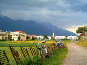 Landscape with mountains against a cloudy sky. There are some buildings in the distance. A slanted brown fence sits on a bed of vibrant green grass. Bikes rest against the fence beside a biking trail.