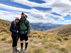 Katelyn Brown-Gomez (right) with her husband John (left) in New Zealand. They are standing on dry grass in front of a mountain scape. They are wearing hiking gear. Brown-Gomez is holding a hiking stick.