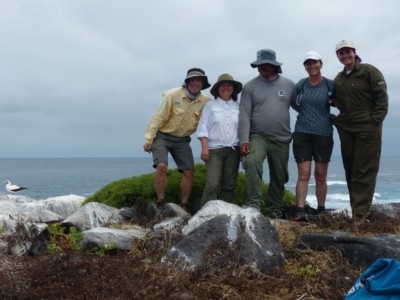 Five scientists stand together, shoulder to shoulder, on a rock in front of the ocean. The sky is cloudy and gray. A white bird stands near them to the left.