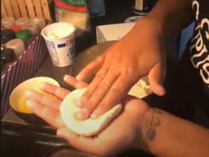 A round piece of dough since in the palm of her left hand as she flatens the dough with the fingers of her right hand. There is a tub of sour cream in the background.