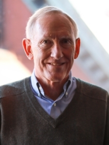Headshot of Ken Brill. He is in a sunlit room and wearing a gray sweater over a blue collared shirt.