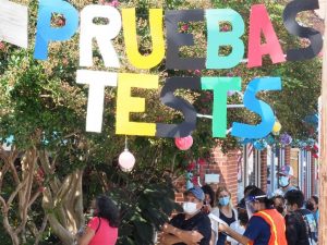 Large hanging letters in alternating colors of blue, white, yellow, green, red and black spelling "pruebas" in Spanish and "tests" in English. People standing in line below the sign. Outdoors.
