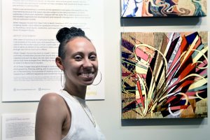 A woman standing in front of two pieces of canvases that display collage art. There is a text panel hanging beside the two pieces.