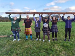 6 students outside wearing kilts. Their arms are above their heads propping up a wooden pole.
