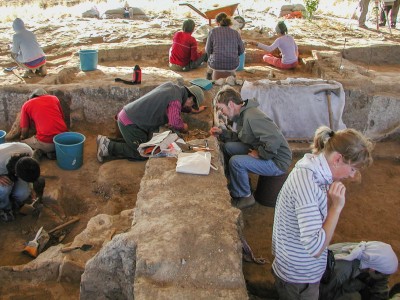People in background and foreground bent out, looking down and digging in the dirt at an archaeological site.