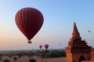 Hot air balloons in the air. A person sits atop the edge of a Burmese temple.