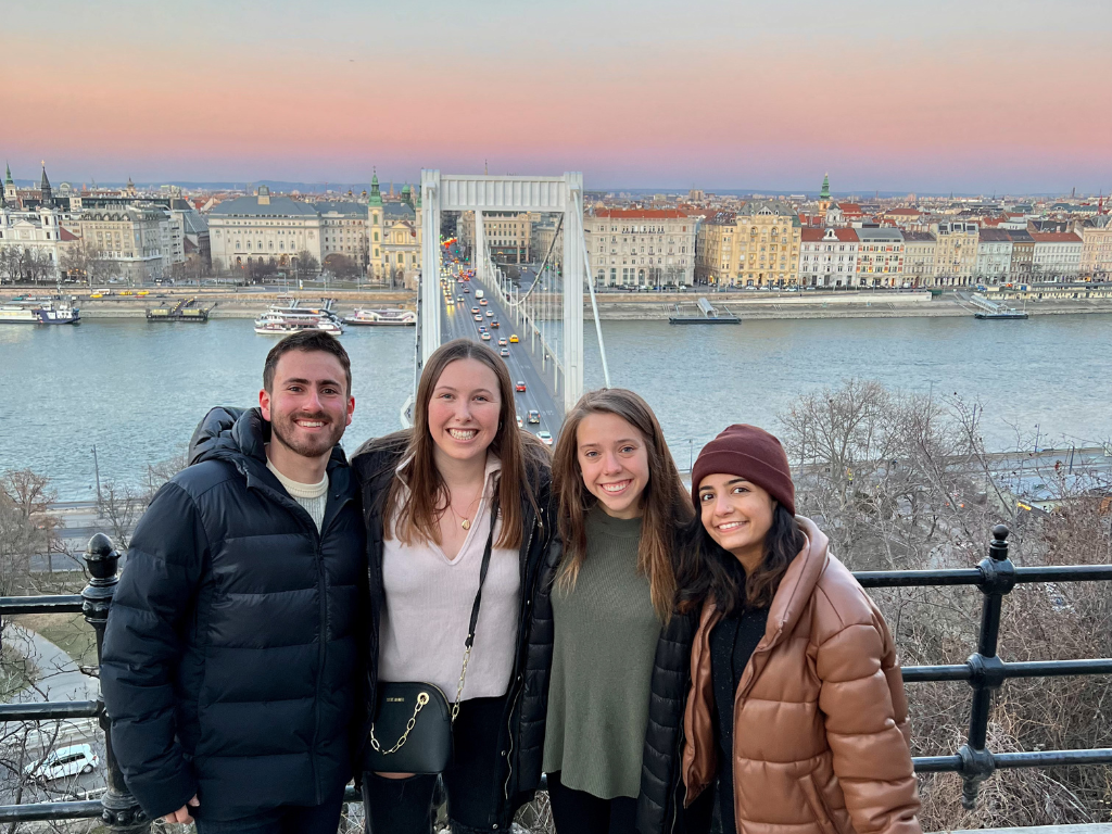 Four students standing together, smiling. There are three women and one man. In the background, a bride with cars moving on it. And a sunset in the sky, with blue, orange and teal hues. There's a body of water under the bridge.