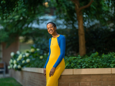 The woman wears a vibrant yellow and blue dress. She is smiling, her posture slouched, which shows an ease about her.