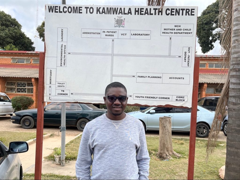 Author standing in front of a sign that says "welcome to Kamwala Heath Center"