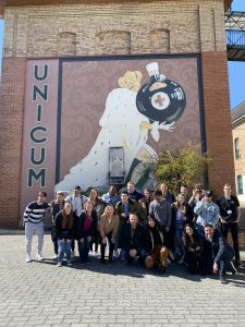 A large group of students standing in front of a mural with a king on it. The sign reads "UNICUM"