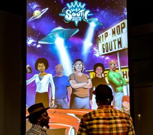 Two people standing in front of an art piece. The art is a painting of people standing under a starry sky with UFO spaceships above. There's a bill board that reads "Hip-Hop South"