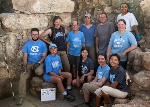 10 UNC students stand in front of a rock quarry. 5 students stand in the back and 5 sit or kneel on rocks in the front.
