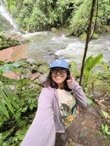 Girl takes a selfie. Smiles and shows a "thumbs up". Behind her, a river flows and there is green foliage.