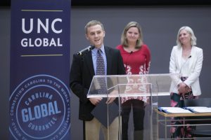 Man standing at a podium, speaking into a microphone. Banner in the background indicates this is a UNC Global event.