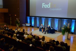 two people sitting on a stage. Audience is in the frame. Behind the speakers, a projector screen with the FedEx Corporation logo.