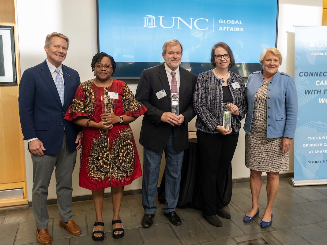 Group of people stand together. Three of them are holding glass trophies. Behind them, a tv screen reads "UNC Global Affairs"