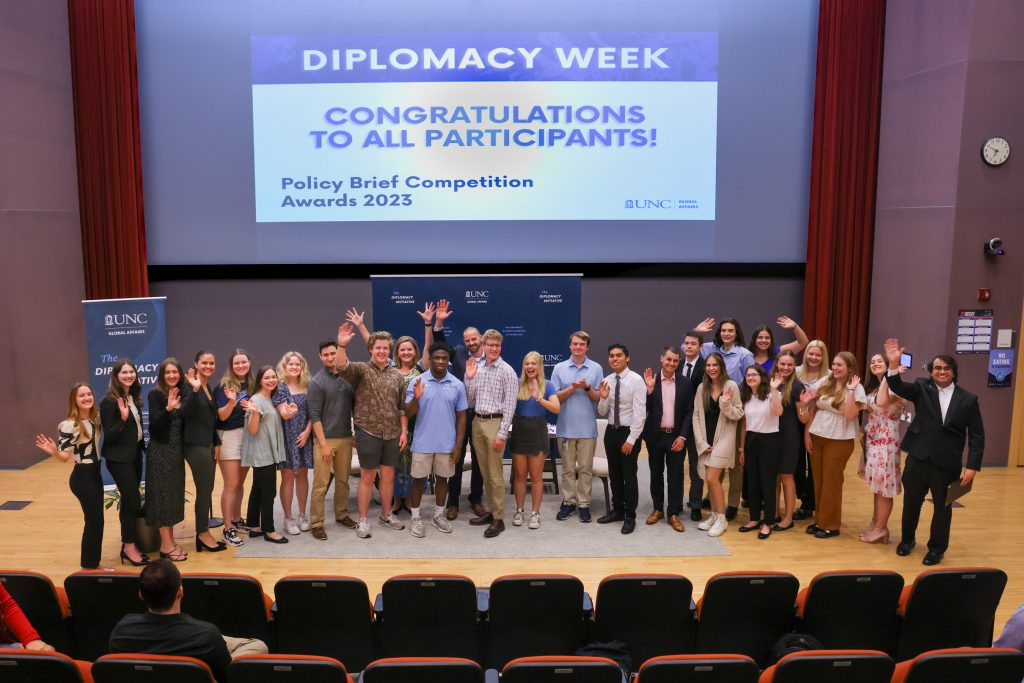 Twenty-four students, dressed in business casual, stand at the front of an auditorium. The backdrop features a projector screen that has a slide reading "Diplomacy Week: Congratulations to all participants!". The students wave and smile enthusiastically at the camera.