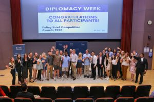 A group of people posing for a photo in front of a large screen in an auditorium. The group is standing close together and smiling for the camera. The screen behind them is displaying a slide with text "Diplomacy Week: Congratulations to all participants!" The auditorium seating is visible in the foreground. The setting is professional.