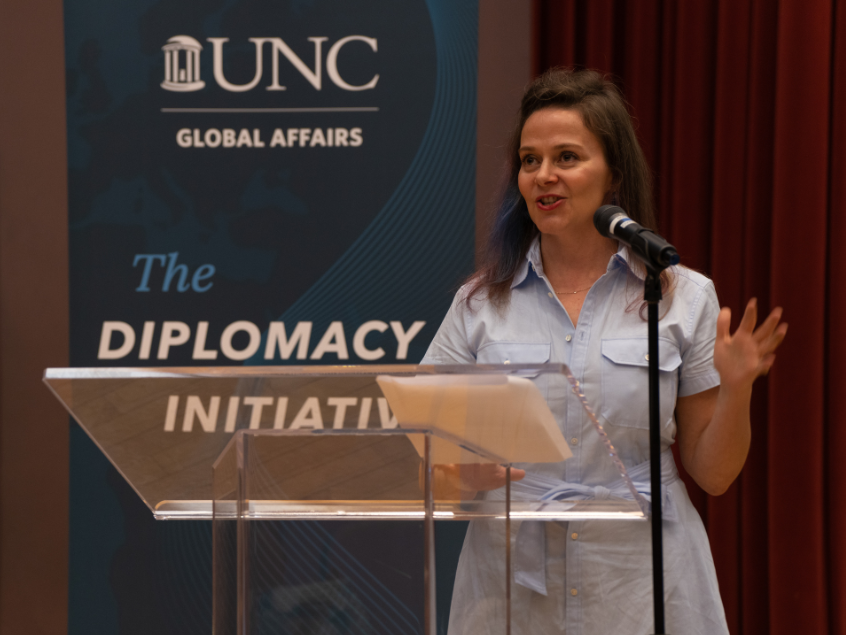 Woman speaking into a microphone at a podium. Behind her, a banner that reads "UNC Global Affairs The Diplomacy Initiative"